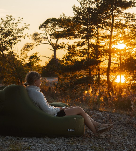 Kvinna sitter i saccosäck på marken i naturen och kollar ut över en solnedgång